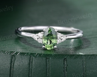 May include: A silver ring with a green oval gemstone surrounded by smaller white gemstones. The ring is on a green surface.