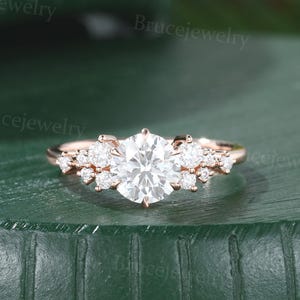 May include: Rose gold engagement ring with a large, round, clear central stone and smaller accent stones. The ring has a delicate rose gold band and an intricate setting, highlighting the sparkle of the gemstones. The ring is displayed against a green background.