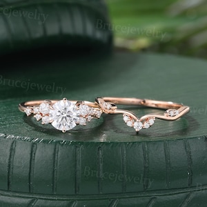May include: Two rose gold rings. The engagement ring has a large, round, clear gemstone with smaller stones around it. The wedding band is a curved design with small gemstones. The rings are on a green surface.