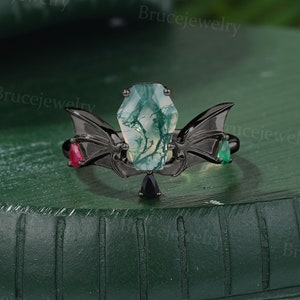 May include: Black ring featuring a green agate stone at its centre, flanked by two black bat wings with red and green gemstones.