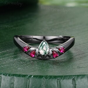 May include: A black metal ring featuring a central pear-shaped moss agate stone flanked by four smaller pear-shaped red stones. The band has a V-shaped contour and a smooth, dark finish, showcasing a distinct contrast between the dark metal and stones.