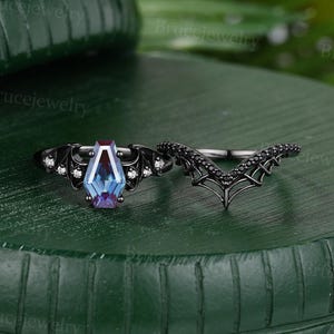 May include: A black metal ring set featuring a coffin-shaped color-changing stone flanked by bat-wing motifs with small clear stones, paired with a matching V-shaped band adorned with tiny black stones arranged in a web-like pattern.