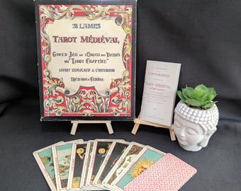 May include: A tarot card set with a box and booklet, displayed on small easels. The box reads "78 LAMES TAROT MÉDIÉVAL" in red and gold lettering. Several tarot cards are fanned out, with a Buddha head planter and succulent to the right.