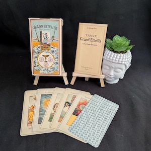 Può includere: Un mazzo di tarocchi Grand Etteilla, un libretto di istruzioni beige intitolato Le Livre de Thot Tarot Grand Etteilla e cinque carte dei tarocchi esposte su piccoli cavalletti di legno. Un vaso bianco a forma di testa di Buddha con una succulenta.