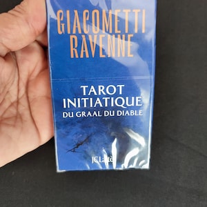 Peut inclure: Un jeu de cartes de tarot bleu intitulé "Giacometti Ravenne" avec des lettres orange. Le jeu est enveloppé dans du plastique transparent. Le texte "Tarot Initiatique Du Graal Du Diable" est également visible.