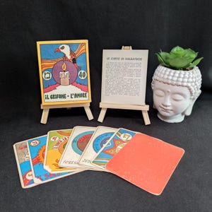 May include: A collection of vintage tarot cards and decorative items. The cards feature colorful illustrations and text, including "IL GRIFONE - L'AMORE." A small Buddha head planter holds a succulent. The cards are displayed on miniature wooden easels.