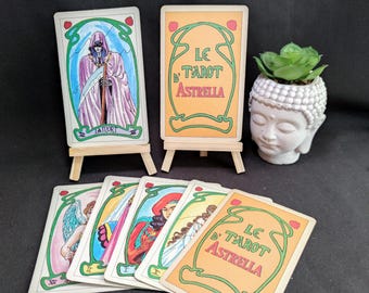 May include: A tarot card deck titled Le Tarot Astrella featuring illustrated cards with green borders and red accents. Two cards are displayed on small wooden easels, while others are fanned out below. A white Buddha head planter with a green succulent sits nearby.