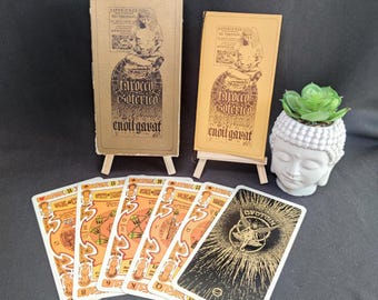 May include: A deck of tarot cards featuring orange and black designs, displayed with two rectangular boxes on small wooden easels and a white ceramic head-shaped planter containing a green succulent. The boxes feature the text Tarocco Esoterico.