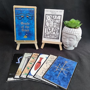 Op de afbeelding: Een Oracle of the Triad kaartendeck met een blauwe doos met een oogillustratie en een wit instructieboekje op houten ezels. Een witte Boeddha-hoofd plantenbak met een groene vetplant staat ernaast, met verschillende kaarten uitgewaaierd eronder.