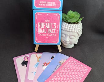 May include: A blue and pink RuPaul's Drag Race tarot card deck box with the text Gaze into your future with this sickening deck! displayed on the lid. Four illustrated cards are fanned out in front, with one pink patterned card face down.