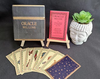 May include: A deck of Oracle Belline cards with a dark gray box featuring gold lettering, a red instructional booklet on a small wooden easel, and a white Buddha-shaped planter with a green succulent, alongside several illustrated cards.