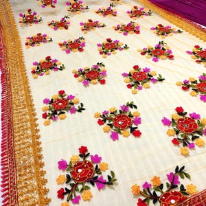 May include: Cream-colored fabric with embroidered floral patterns in red, pink, and yellow. The design features repeating clusters of flowers with mirror accents. The edges are trimmed with gold and red decorative borders, creating a vibrant and detailed textile.