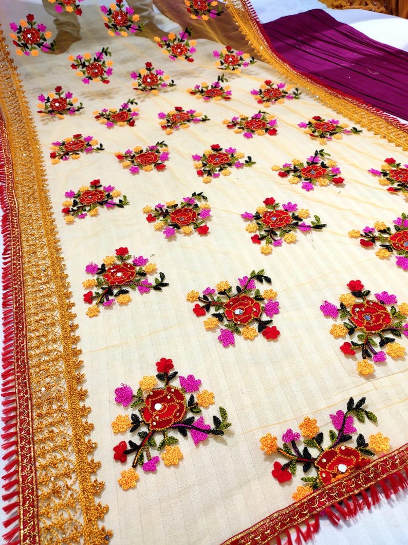 May include: Cream-colored fabric with embroidered floral patterns in red, pink, and yellow. The design features repeating clusters of flowers with mirror accents. The edges are trimmed with gold and red decorative borders, creating a vibrant and detailed textile.