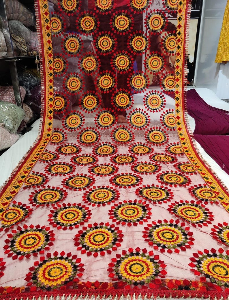 May include: A sheer, red shawl with a repeating pattern of embroidered floral medallions in yellow, red, and black. The edges are trimmed with a yellow and red geometric border and a red fringe.