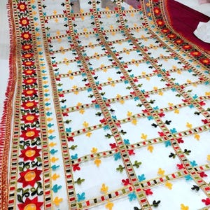 May include: A sheer white fabric with a grid pattern, adorned with colorful floral and geometric embroidery. The edges are trimmed with a red and gold border, and the fabric has a fringed edge.