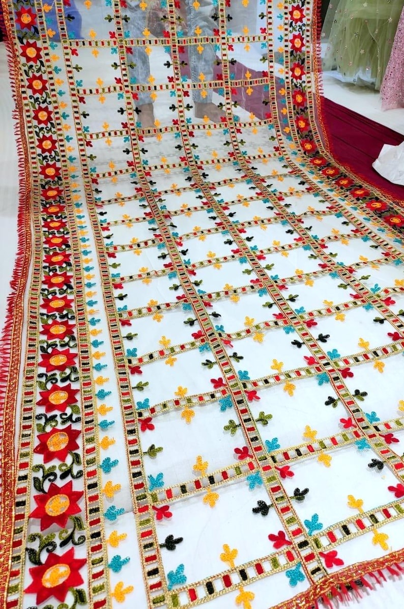May include: A sheer white fabric with a grid pattern, adorned with colorful floral and geometric embroidery. The edges are trimmed with a red and gold border, and the fabric has a fringed edge.