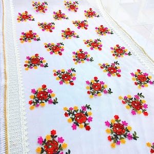 May include: White fabric with embroidered floral patterns in red, pink, yellow, and green. The edges are trimmed with lace and gold accents. The floral design is repeated across the fabric.