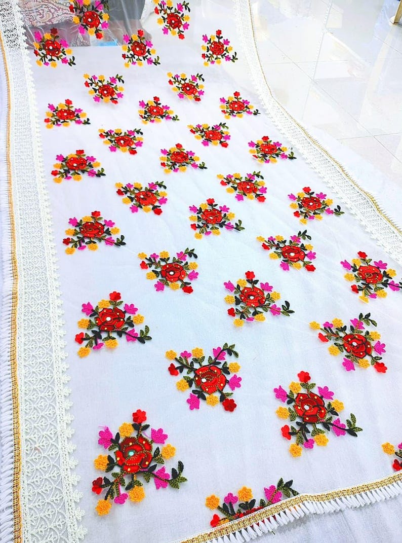 May include: White fabric with embroidered floral patterns in red, pink, yellow, and green. The edges are trimmed with lace and gold accents. The floral design is repeated across the fabric.