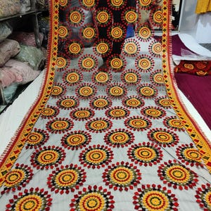 May include: A sheer, gray fabric with a repeating pattern of red, yellow, and black circular designs. The edges are trimmed with a decorative border in red, yellow, and black. The fabric appears to be a shawl or scarf.