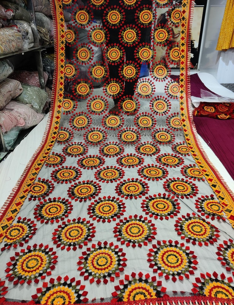 May include: A sheer, gray fabric with a repeating pattern of red, yellow, and black circular designs. The edges are trimmed with a decorative border in red, yellow, and black. The fabric appears to be a shawl or scarf.