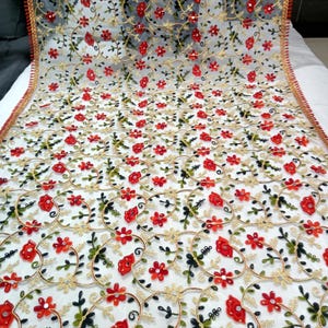 May include: A sheer white fabric with intricate embroidery. The design features red flowers, green leaves, and gold-colored circular patterns. The edges are trimmed with a red and gold border, creating a delicate and ornate textile.