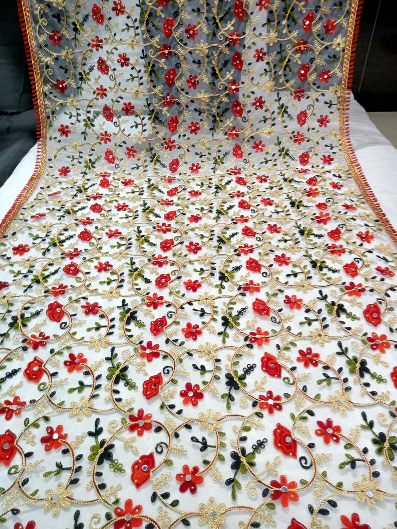 May include: A sheer white fabric with intricate embroidery. The design features red flowers, green leaves, and gold-colored circular patterns. The edges are trimmed with a red and gold border, creating a delicate and ornate textile.