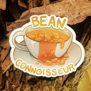 May include: A die-cut sticker featuring a white teacup and saucer filled with orange baked beans. The beans are overflowing down the side of the cup. The text "BEAN CONNOISSEUR" is printed in yellow letters above and below the teacup illustration.