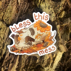 May include: A white ceramic mug with a black handle and the text "But First, Coffee" in a bold, black font. The mug is set against a blurred background, suggesting a cozy setting.