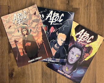 Könnte beinhalten: Drei Comics mit dem Titel "A.P.O.C" werden auf einer Holzoberfläche präsentiert. Die Cover zeigen Illustrationen von Charakteren und Szenen in verschiedenen Farben, darunter Orange, Blau und Schwarz. Der Name des Autors, Hannah Spikings, ist auf jedem Cover aufgedruckt.