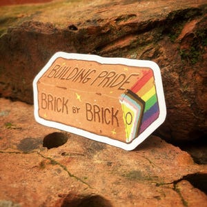 May include: A rectangular sticker shaped like a brown brick with the text "BUILDING PRIDE BRICK BY BRICK" in brown lettering. The right side of the brick features a vertical rainbow-coloured pride flag design with additional light blue, pink, white, brown, and black stripes.