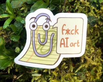 May include: A sticker featuring a cartoon paperclip with googly eyes holding a speech bubble. The speech bubble contains the text "F*ck AI art" in a handwritten style. The sticker is set against a background of green foliage.