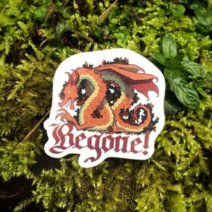 May include: A sticker depicting an orange dragon with red wings and green detailing. The dragon is set against a white background, with the word "Begone!" in a decorative font below. The sticker is placed on a bed of green moss.