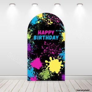 May include: A black arch-shaped party backdrop with colorful paint splatters in pink, yellow, blue, and green. The words "HAPPY BIRTHDAY" are in bright pink and blue. The backdrop is designed for a birthday celebration.