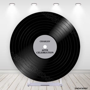 May include: A large black vinyl record with the text "CHARLES' 65TH CELEBRATION" in a gray center label. The record is displayed on a silver stand against a white wall with spotlights. The stand is not included.