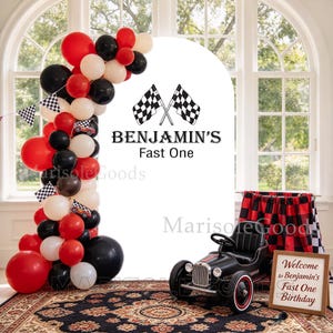 May include: A white arched backdrop featuring two crossed checkered flags and the text "BENJAMIN'S Fast One". A balloon garland in red, black, and white surrounds the arch. A black toy race car and a sign reading "Welcome to Benjamin's Fast One Birthday" are placed in front.