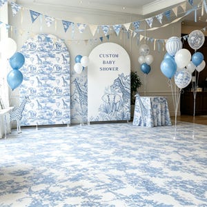 Può includere: Un allestimento per baby shower con due fondali ad arco decorati con stampe di animali blu e bianche, uno dei quali riporta la scritta CUSTOM BABY SHOWER. Il set include palloncini blu e bianchi, una tovaglia coordinata e festoni.