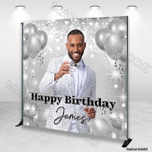 Puede incluir: Un telón de fondo de cumpleaños plateado y blanco con un hombre con un traje texturizado sosteniendo un cóctel. El telón de fondo incluye globos plateados, confeti y el texto "Happy Birthday James."