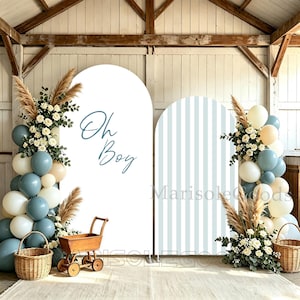 Könnte beinhalten: Zwei bogenförmige Hintergrundpaneele, eines in Weiß mit dem Schriftzug "Oh Boy" in blauer Schreibschrift und eines mit hellblauen vertikalen Streifen. Beide sind mit Ballongirlanden in Blau, Creme und Weiß, Pampasgras und einem Holzwagen dekoriert.