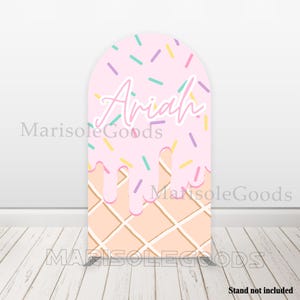 May include: Pink and white ice cream cone shaped backdrop with the name Ariah in pink lettering and colorful sprinkles.