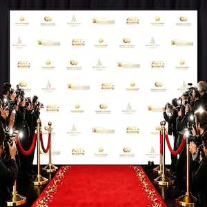 Puede incluir: Una alfombra roja conduce a un telón de fondo blanco con logotipos dorados, flanqueado por cuerdas de terciopelo y postes. Fotógrafos con cámaras y flashes se alinean a los lados, creando una escena que recuerda a un estreno de cine o una ceremonia de premios.
