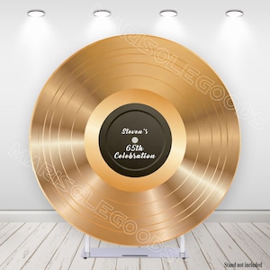 May include: A large, gold-colored vinyl record with a black center label. The label reads "Steven's 65th Celebration" in white text. The record is displayed on a silver stand, and the background is a white wall and a wooden floor.