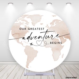Puede incluir: Un telón de fondo redondo blanco con un mapa del mundo en tonos marrones apagados. El texto "OUR GREATEST adventure BEGINS" está escrito sobre el mapa, con un corazón y un avión. El soporte no está incluido.