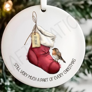 May include: A white Christmas tree ornament with a red stocking design and a tag that says "GEORGE". A small bird is perched on the stocking. The ornament has the text "STILL VERY MUCH A PART OF EVERY CHRISTMAS" in a circular pattern.