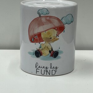 May include: White ceramic money box featuring a cartoon duck wearing a red umbrella and boots. The money box has the words "Rainy day FUND" written on it.