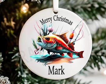 Puede incluir: Adorno navideño blanco con un pez colorido, cañas de pescar y el texto «Merry Christmas Mark». El pez es rojo, naranja y azul. El adorno tiene una cinta blanca para colgar. Decoración festiva.