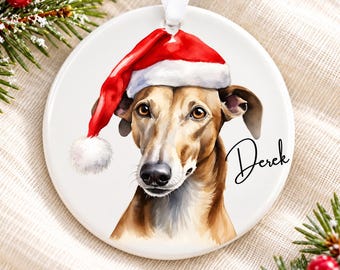 May include: A round white ceramic ornament featuring a watercolor illustration of a tan greyhound wearing a red and white Santa hat. The name Derek is printed in black script on the right side. A white ribbon is attached to the top for hanging.