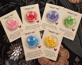 Op de afbeelding: Een verzameling Pocket Hugs, elk met een helder, cirkelvormig ontwerp met een lachend gezicht gevuld met kleurrijke glitter. De kaarten lezen "Pocket Hug" en bevatten een bericht over veilig bewaren.