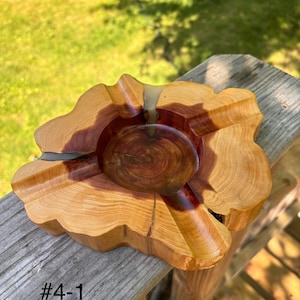 May include: A handcrafted wooden ashtray with a natural, organic shape. The ashtray features a central bowl with a dark, reddish-brown hue, surrounded by a lighter, golden-brown wood grain. The item is marked with the text "#4-1".