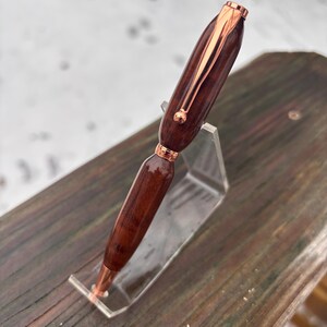 May include: A handcrafted wooden pen with copper-coloured accents. The pen features a dark brown, polished wooden barrel and a copper-coloured clip and tip. The pen is displayed on a clear acrylic stand, showcasing its elegant design.