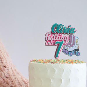 May include: A teal, pink and white cake topper with the text "Olivia Rolling into 7" and a cartoon image of roller skates.